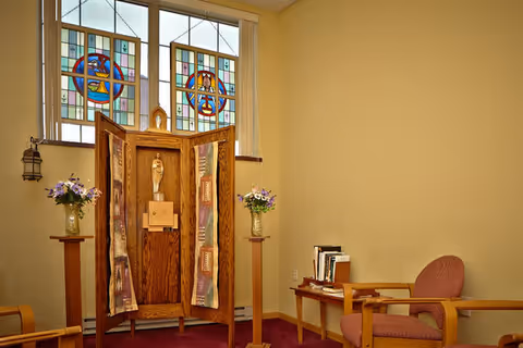 Small chapel-style interior with a wooden shrine and statue beneath stained-glass windows, flanked by chairs and flower arrangements.