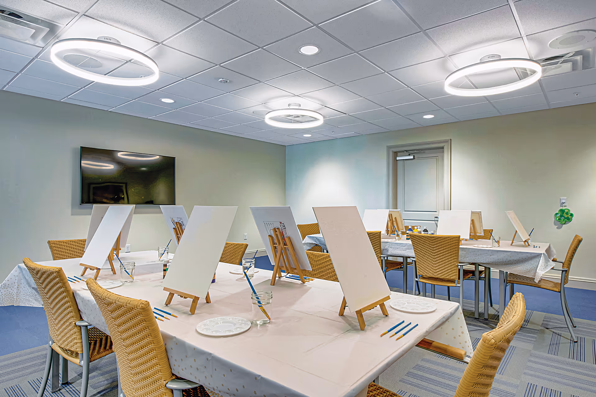 Bright indoor activity room with tables arranged for painting, each set with easels, blank canvases, brushes, and chairs under circular ceiling lights.