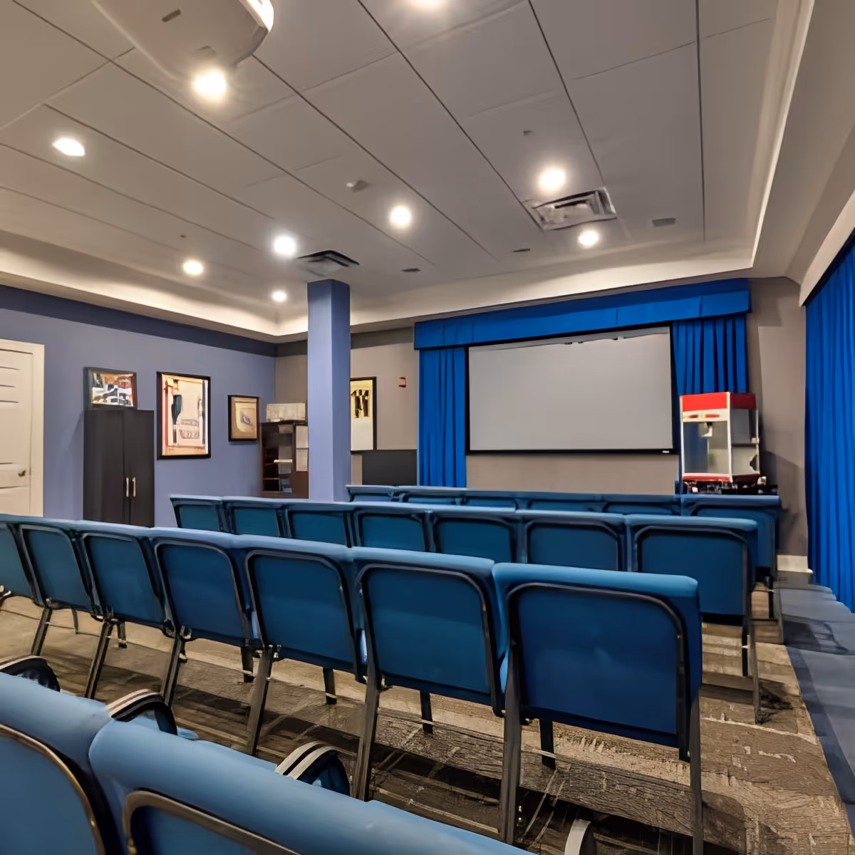 Interior view of a small theater or presentation room with rows of blue chairs facing a large white projection screen framed by blue curtains. The room has a blue and gray color scheme with framed artwork on the walls and a popcorn machine on the right side near the screen.