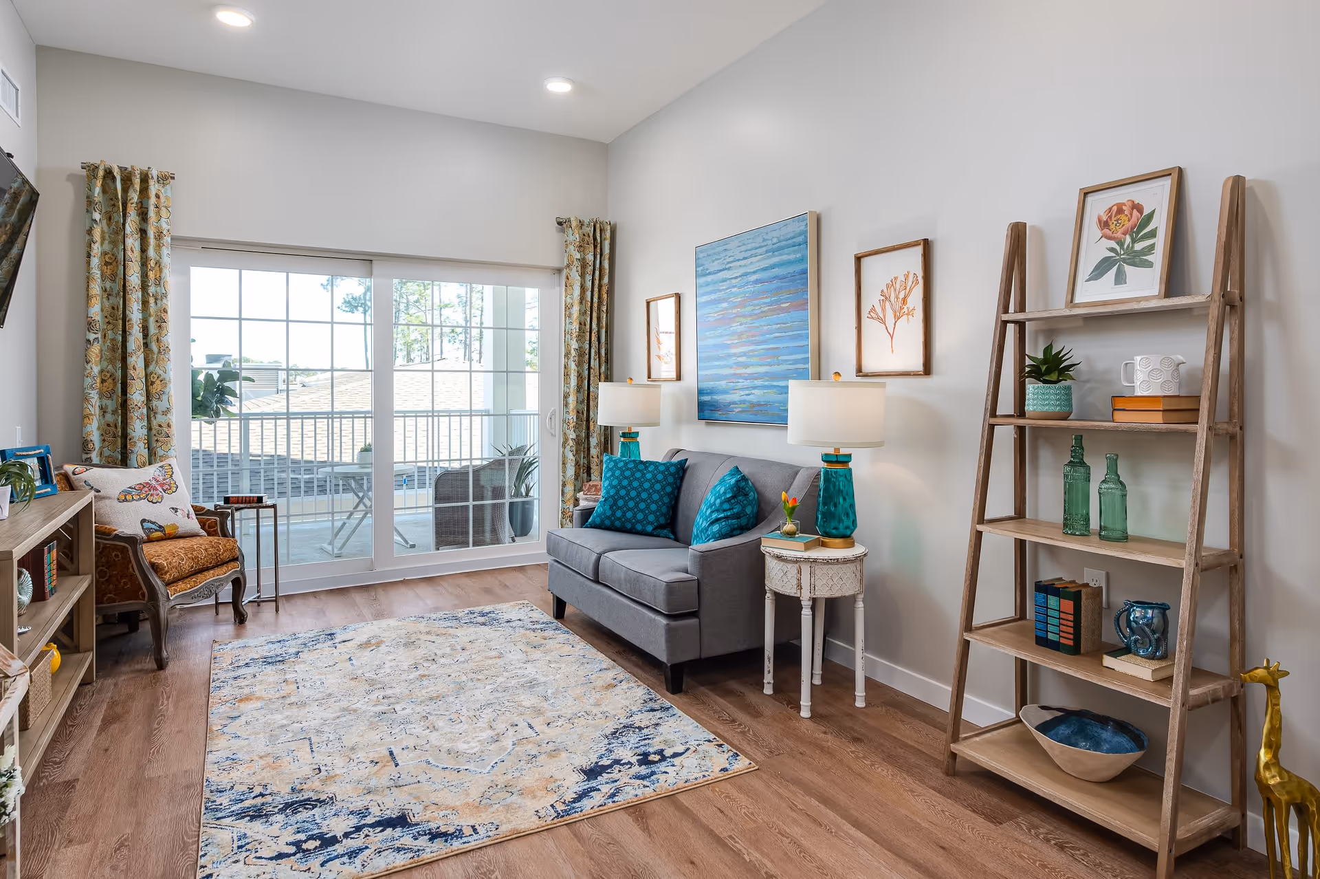 A bright living room with a gray loveseat adorned with teal pillows, two matching teal lamps on white side tables, a wooden ladder shelf with decorative items, a patterned area rug on wooden flooring, a vintage armchair with a butterfly pillow, and large sliding glass doors with floral curtains leading to a balcony.