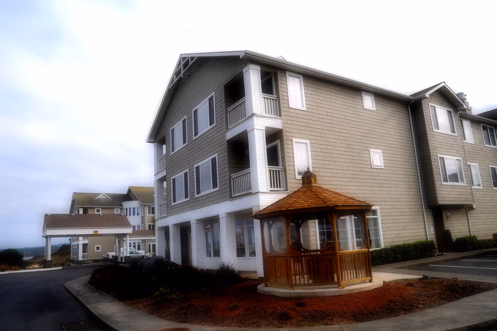 Exterior view of a multi-story senior living facility building with beige siding and white trim. In front of the building is a wooden gazebo with a shingled roof, surrounded by a landscaped area with mulch. A paved driveway and parking area are visible, along with another building in the background under a cloudy sky.