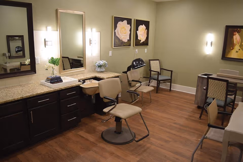 A well-lit salon room with two beige salon chairs in front of a long granite countertop with dark wooden cabinets. There are two large mirrors on the wall, decorative floral paintings, and additional chairs along the walls. The room has wooden flooring and soft green walls.