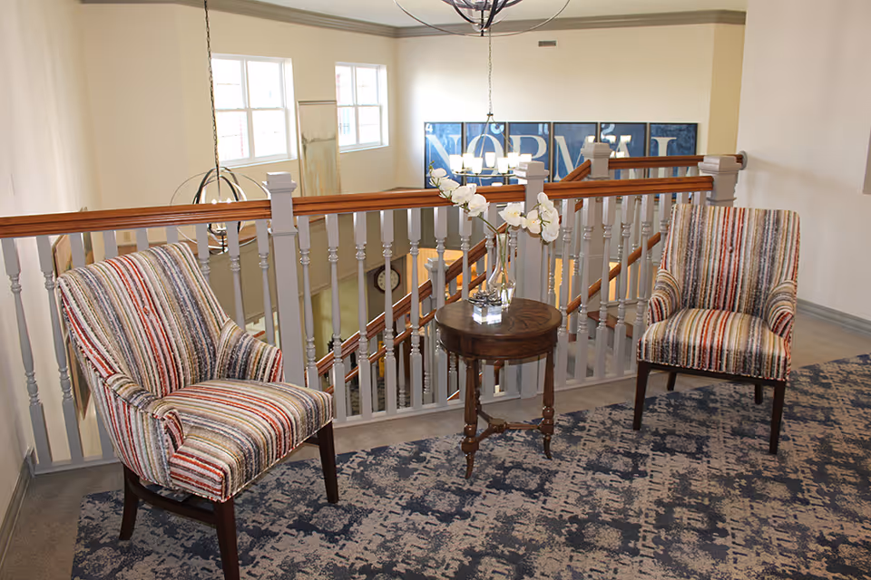 A cozy sitting area with two striped upholstered chairs and a small round wooden table with a glass vase holding white flowers, positioned near a railing overlooking a staircase in a well-lit interior space.