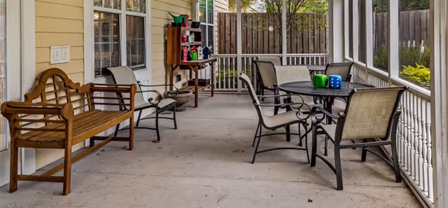 A covered outdoor patio area with a wooden bench, several metal chairs with beige cushions, and a round table with large colorful dice on it. The patio is enclosed with white railings and has a wooden fence and greenery visible in the background.