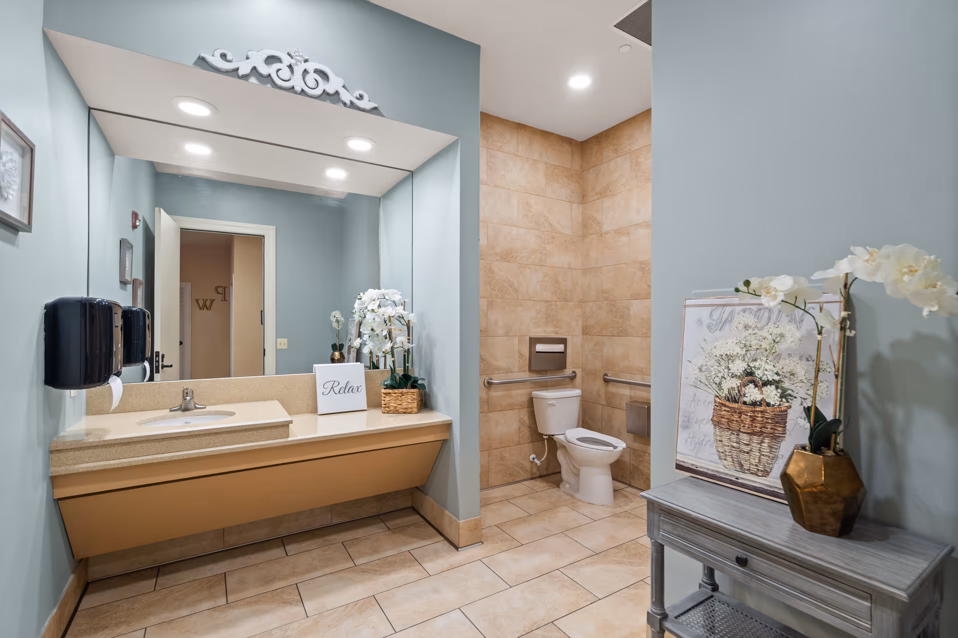 A clean and well-lit bathroom with beige tiled floors and walls. There is a large mirror above a beige countertop with a sink, a soap dispenser, and a decorative sign that says 'Relax'. To the right, there is a toilet with grab bars on the walls for accessibility. A small gray table with a vase of white flowers and a framed floral artwork is positioned against the right wall. The walls are painted a soft blue color.