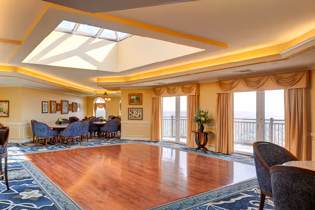 Bright, elegant common room with a skylight, hardwood dance floor, dining tables and chairs, and balcony doors with draped curtains.