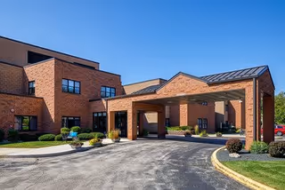 Exterior view of a brick senior living facility with a covered entrance driveway, landscaped bushes, and a clear blue sky.