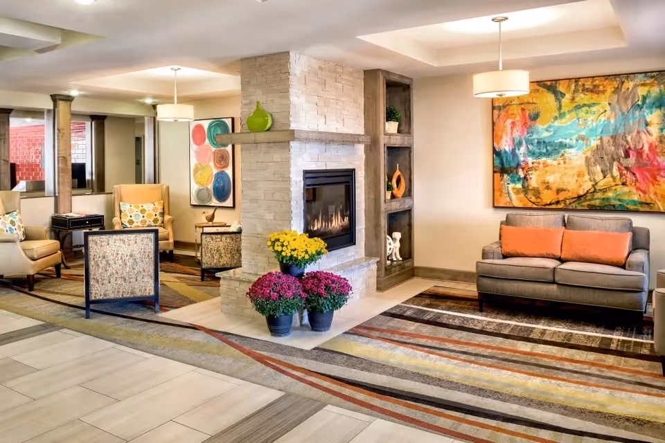 A cozy living room area featuring a modern stone fireplace with a fire burning inside. Around the fireplace are three pots of colorful flowers. The room has a patterned carpet with warm tones, a gray sofa with orange cushions, two armchairs with patterned upholstery, and a large colorful abstract painting on the wall. The space is softly lit with ceiling lights and has decorative shelves with vases and plants.