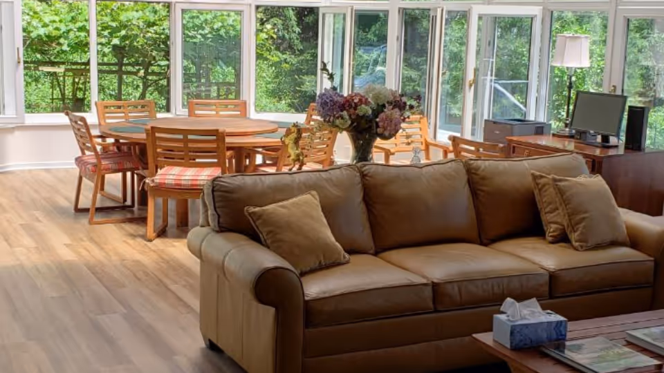 A bright and spacious common area with a tan leather sofa in the foreground, a wooden coffee table with a tissue box and magazines, and a dining table with six wooden chairs with plaid cushions near large windows showing green foliage outside. A desk with a computer and a lamp is visible in the background.