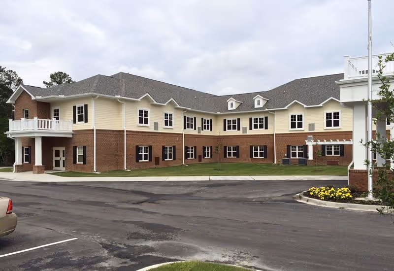 Exterior view of a two-story senior living facility building with a combination of brick and light yellow siding, multiple windows, a balcony, and a parking area in front with a small flower bed near a flagpole.
