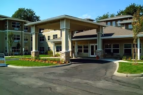 Exterior view of a multi-story senior living facility with a covered entrance driveway, landscaped lawn, and trees under a clear blue sky.