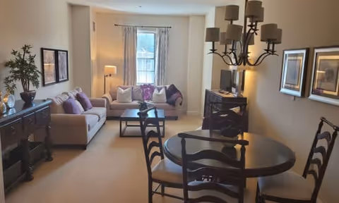 A cozy living room and dining area in a senior living facility featuring a beige sofa with purple and white cushions, a coffee table with a flower arrangement, a round dining table with four chairs, a sideboard with decorative items, framed artwork on the walls, a window with curtains, and a floor lamp in the corner.