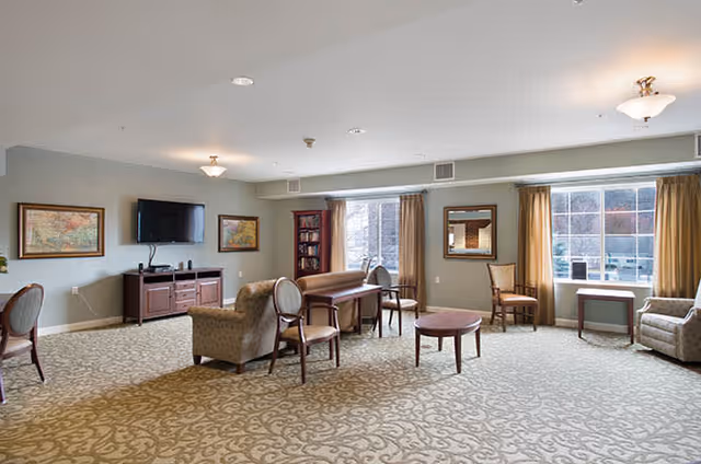 A spacious living room area with patterned carpet, several chairs, a small round table, a TV mounted on the wall above a wooden cabinet, framed maps on the walls, large windows with beige curtains letting in natural light, and a bookshelf in the corner.