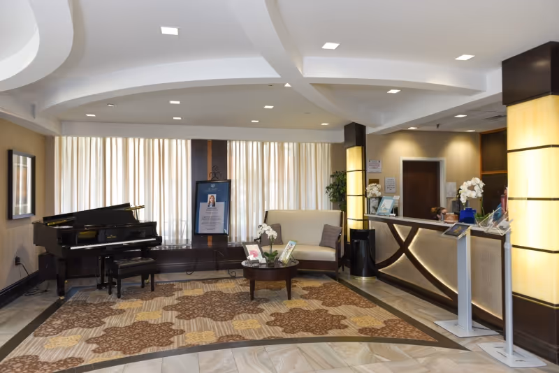 A well-lit reception area with a grand piano on the left, a beige loveseat with a pillow, a round wooden coffee table with decorative items, and a reception desk on the right. The floor has a patterned carpet, and large windows with sheer curtains provide natural light. The ceiling features recessed lighting and a modern design.