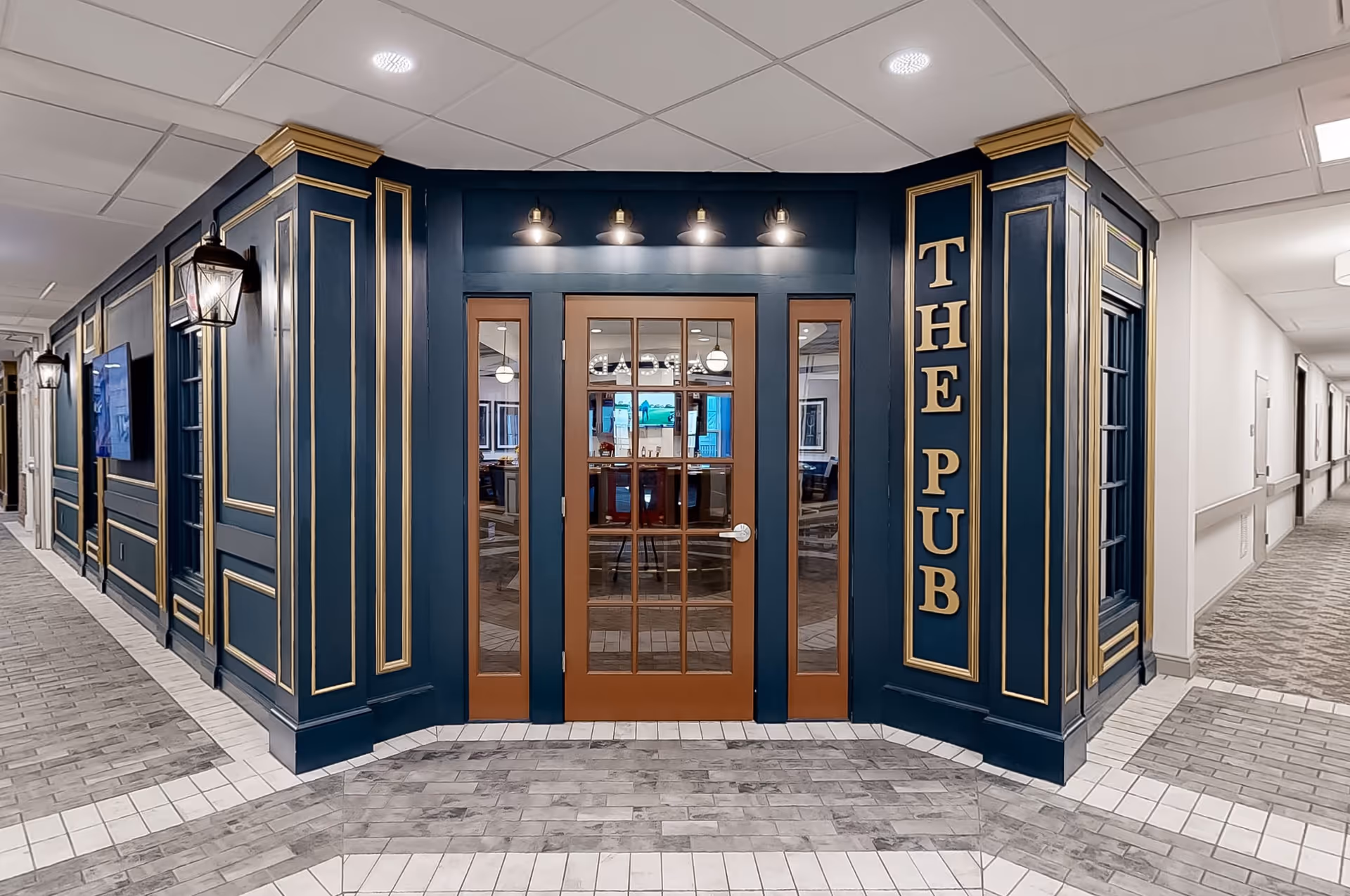 Interior hallway view of a senior living facility featuring an entrance to a room labeled 'THE PUB' with dark blue walls accented with gold trim, glass-paneled double doors, and warm lighting fixtures above the entrance.