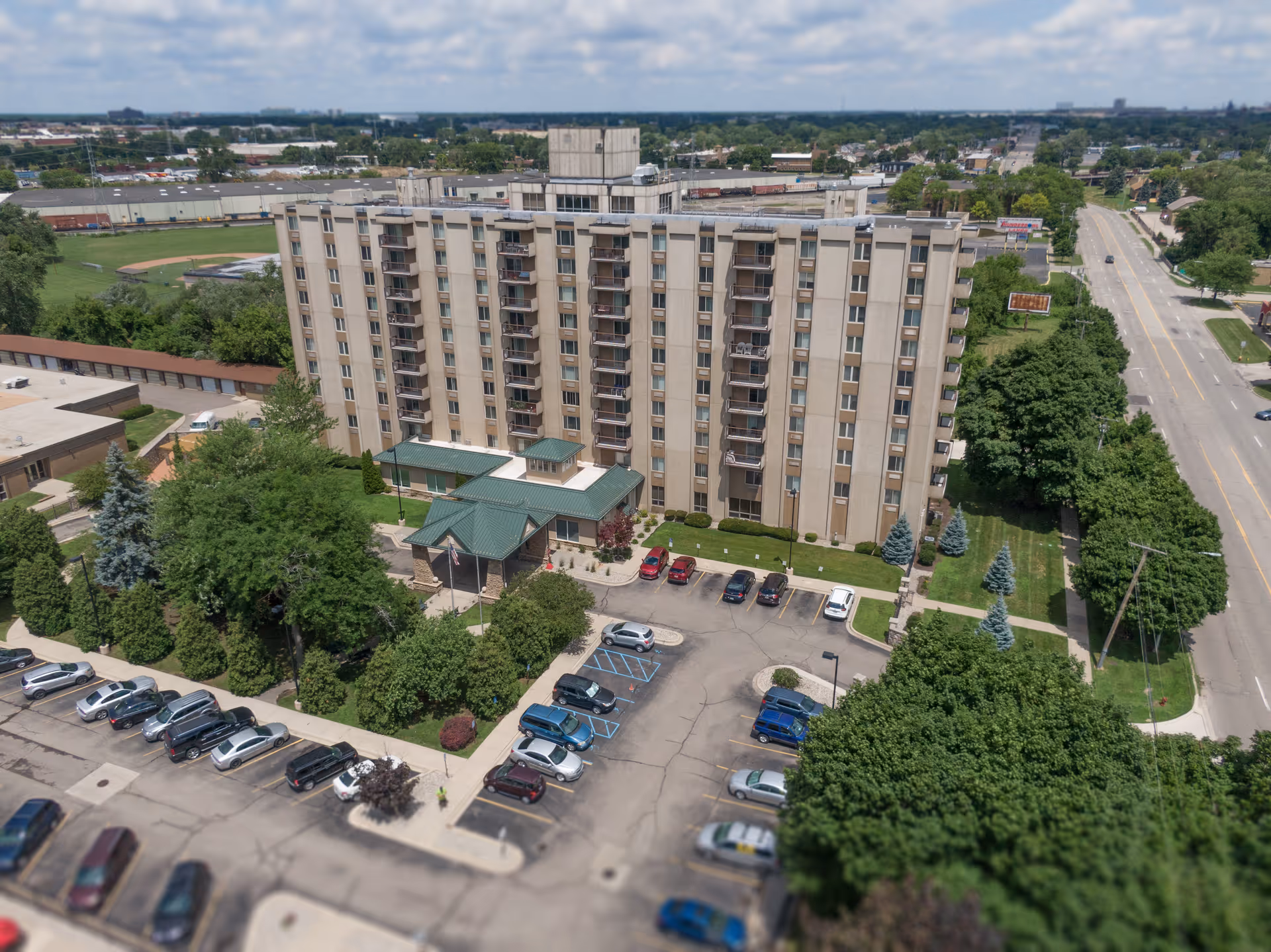 Aerial view of a multi-story senior living facility named Maple Heights Senior Living, surrounded by trees, parking lots with cars, and adjacent roads under a partly cloudy sky.