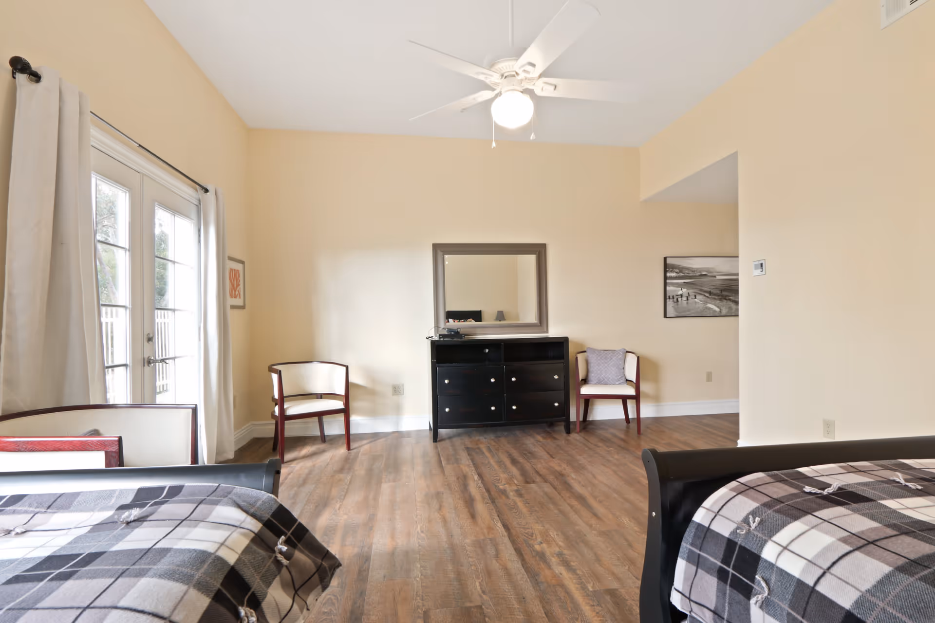 Bright bedroom with two beds, a dresser and mirror, chairs, a ceiling fan, and French doors.