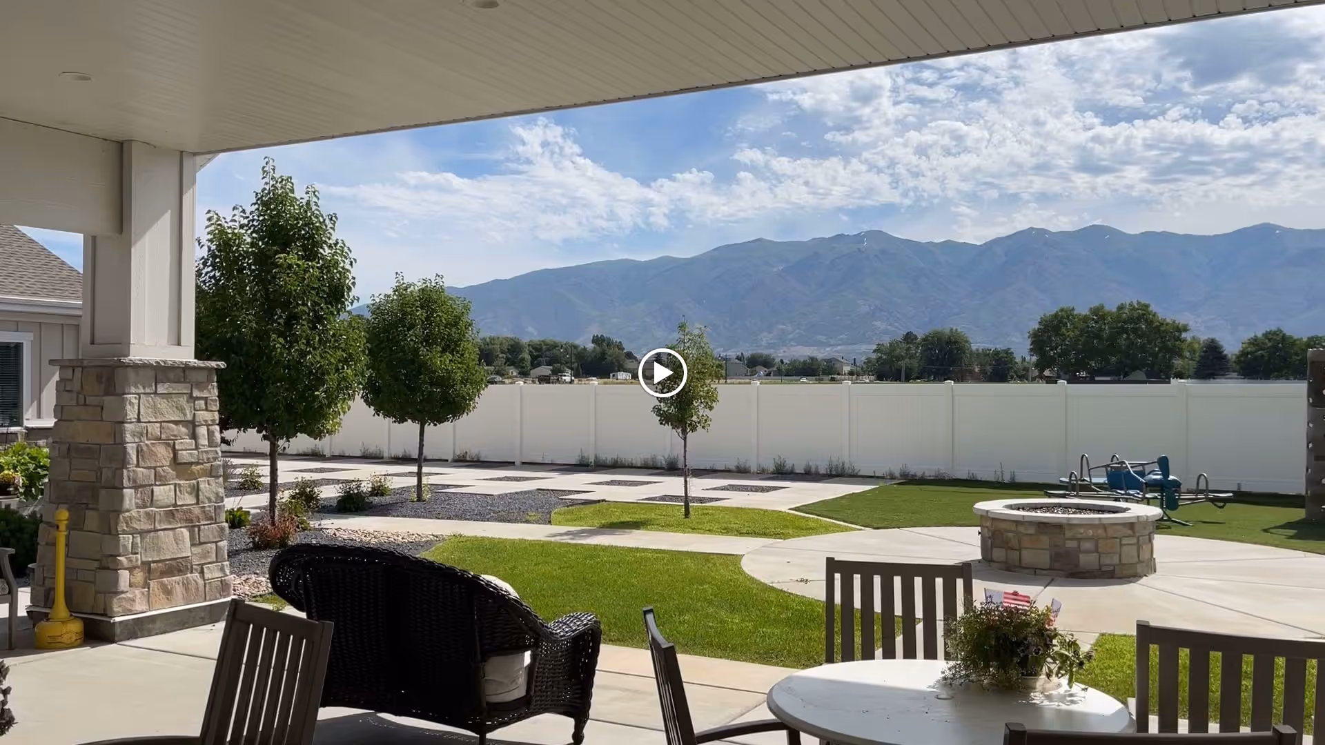 Covered patio area with outdoor seating including a round table with chairs and a wicker loveseat, overlooking a landscaped yard with a fire pit, small trees, and a white fence. Mountains and a partly cloudy sky are visible in the background.