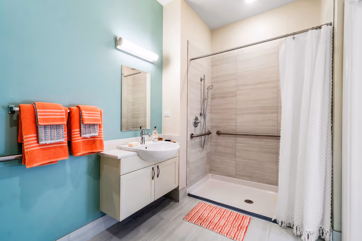 A modern bathroom with a walk-in shower featuring beige tiles and a white shower curtain. There are two orange towels with patterned accents hanging on a towel rack on a teal wall. A white floating vanity with a sink, soap, and a mirror is mounted on the wall beneath a horizontal light fixture. A striped orange and white bath mat is placed in front of the shower.