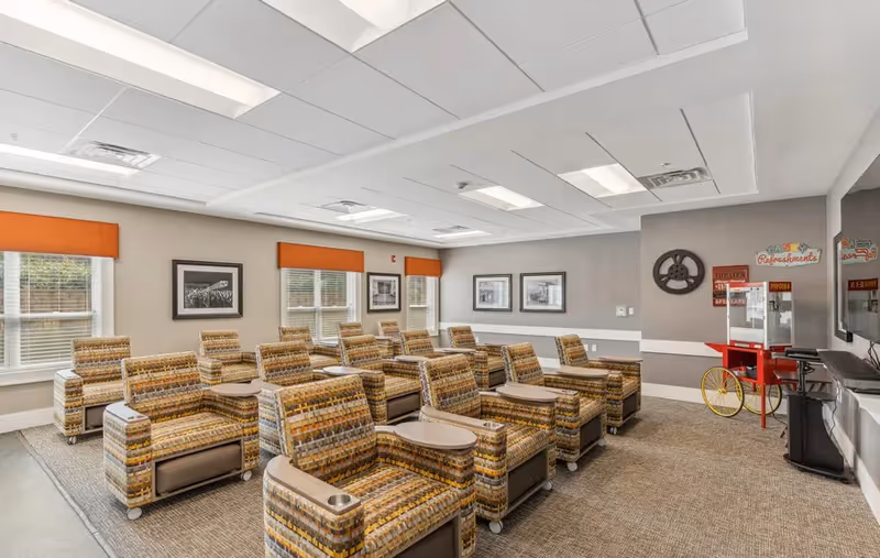 A cozy media room with multiple patterned armchairs arranged in rows facing a large flat-screen TV mounted on the wall. The room has three windows with orange valances, framed black and white photos on the walls, and a vintage-style popcorn machine on a red cart in the corner. The ceiling has recessed lighting and white tiles.
