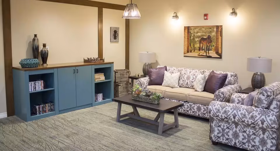 A cozy living room with a patterned sofa and armchair, a wooden coffee table with a plant centerpiece, two table lamps on side tables, a blue cabinet with decorative vases and books, and a framed picture of a horse on the wall.