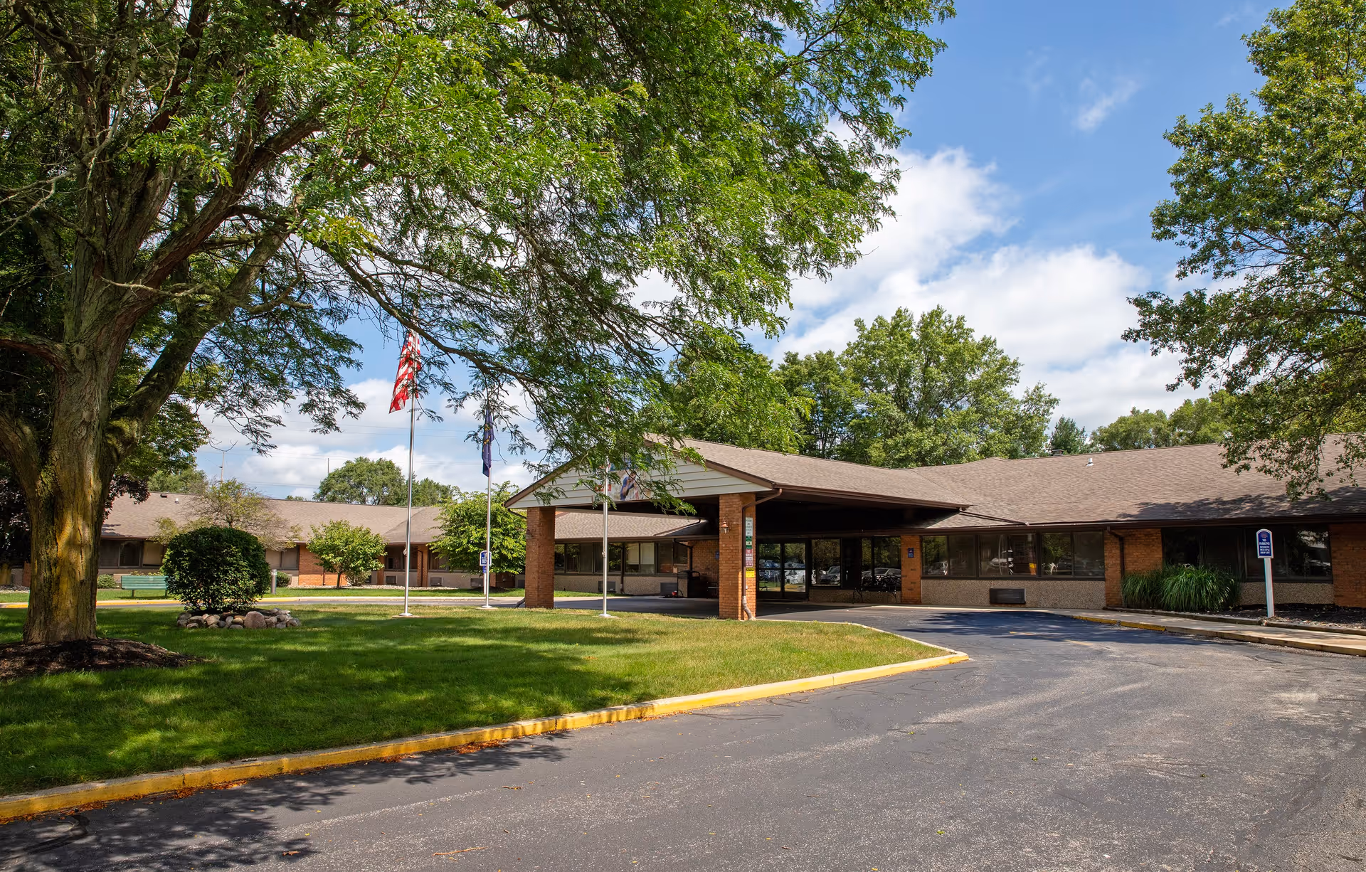 Exterior view of East Lake Nursing and Rehabilitation Center showing a single-story brick building with a covered entrance, surrounded by green trees and a well-maintained lawn under a partly cloudy blue sky.