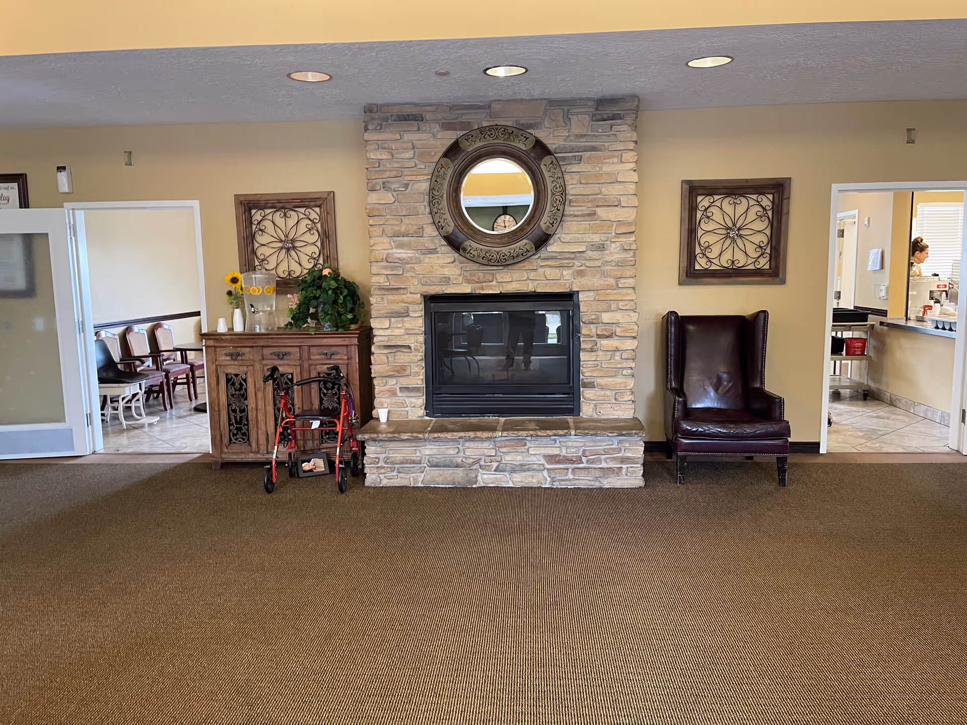 Interior view of a common area in a senior living facility featuring a stone fireplace with a round decorative mirror above it. To the left of the fireplace is a wooden cabinet with a water dispenser and a red walker in front. To the right is a dark leather armchair. Two doorways flank the fireplace, one leading to a room with chairs and the other to a kitchen area where a person is visible working.