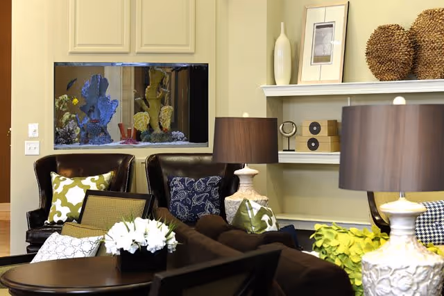 A cozy living room area with dark leather armchairs and a sofa adorned with patterned pillows. A built-in aquarium with colorful coral and fish is set into the wall. The room features a table with a white floral arrangement, two table lamps with dark shades, and decorative items on white shelves including vases, framed art, and woven spheres.