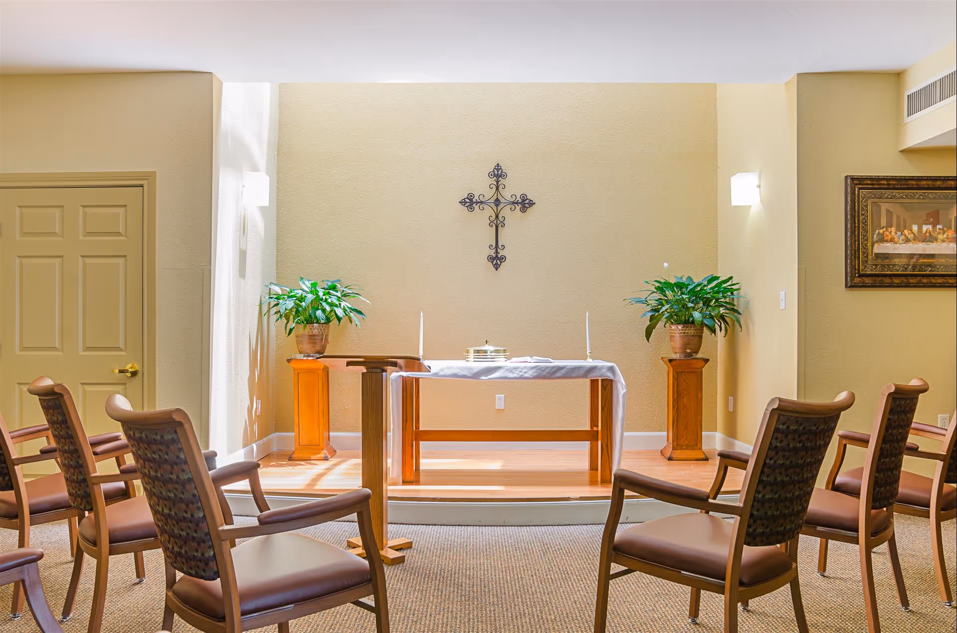 Small chapel-style interior with an altar draped in white, a decorative cross on the wall, potted plants, and rows of chairs.