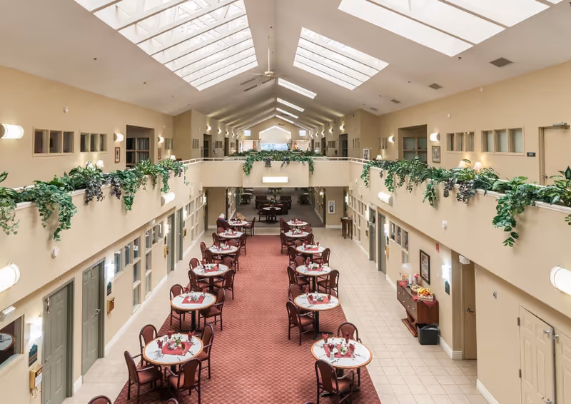 Interior view of a spacious dining area in an assisted living facility with multiple round tables set with flowers and place settings. The room has a high ceiling with skylights, beige walls, and green plants decorating the upper balcony railings. Doors line both sides of the room, and there is a carpeted walkway in the center.