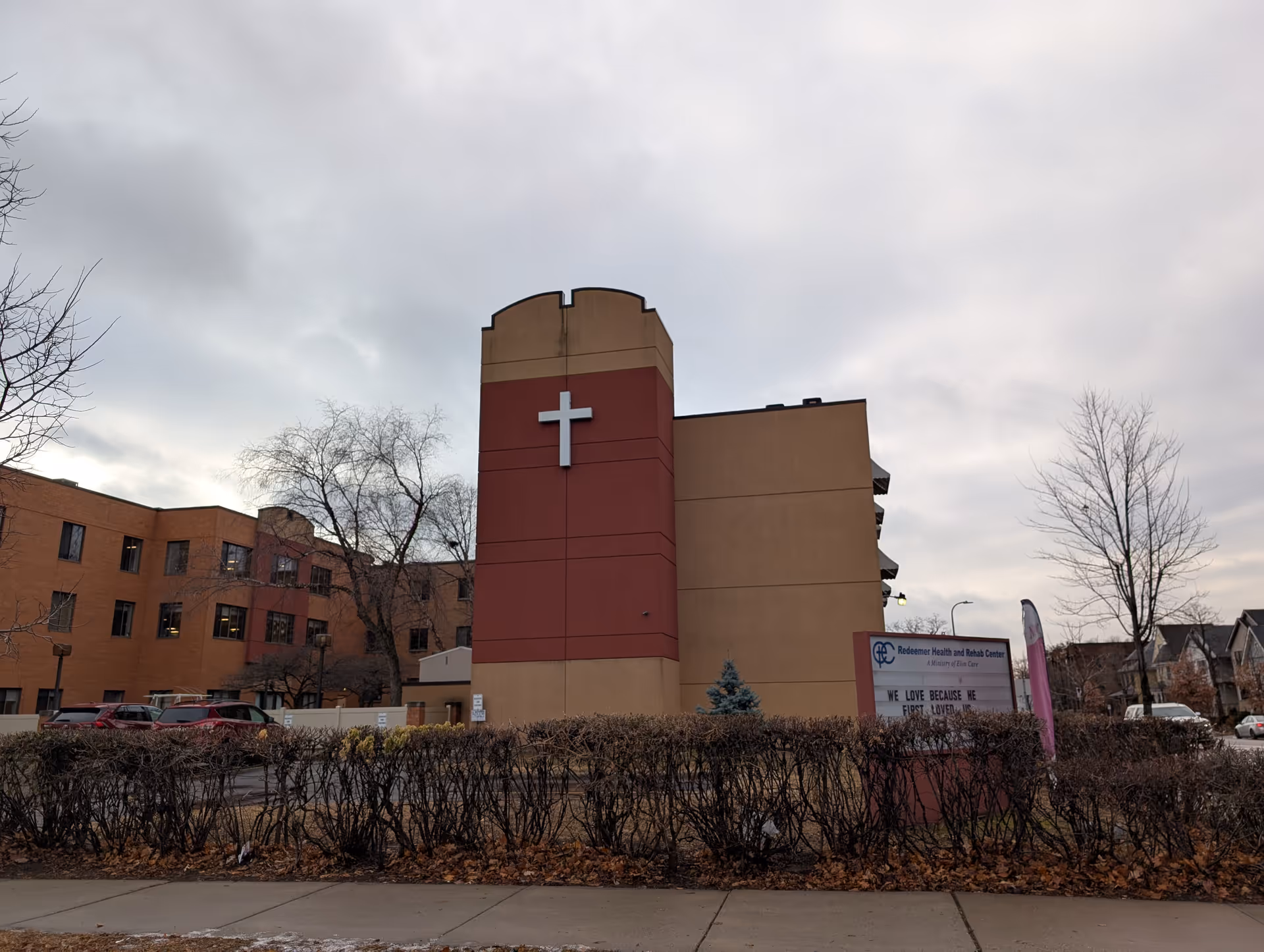 Exterior view of Redeemer Health and Rehab Center building with a large white cross on a red and beige tower. Leafless trees and trimmed bushes surround the building, and a sign in front displays a message. Several cars are parked near the building under a cloudy sky.