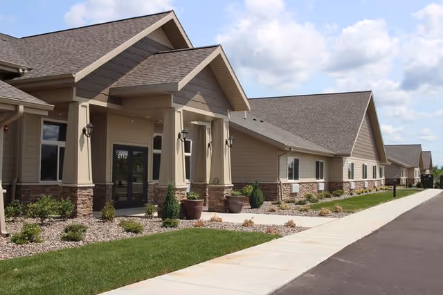 Exterior view of a single-story assisted living facility building with beige siding, stone accents, and a gabled roof. There is a paved walkway and landscaped area with grass, shrubs, and small trees in front of the entrance under a covered porch with columns. The sky is partly cloudy.