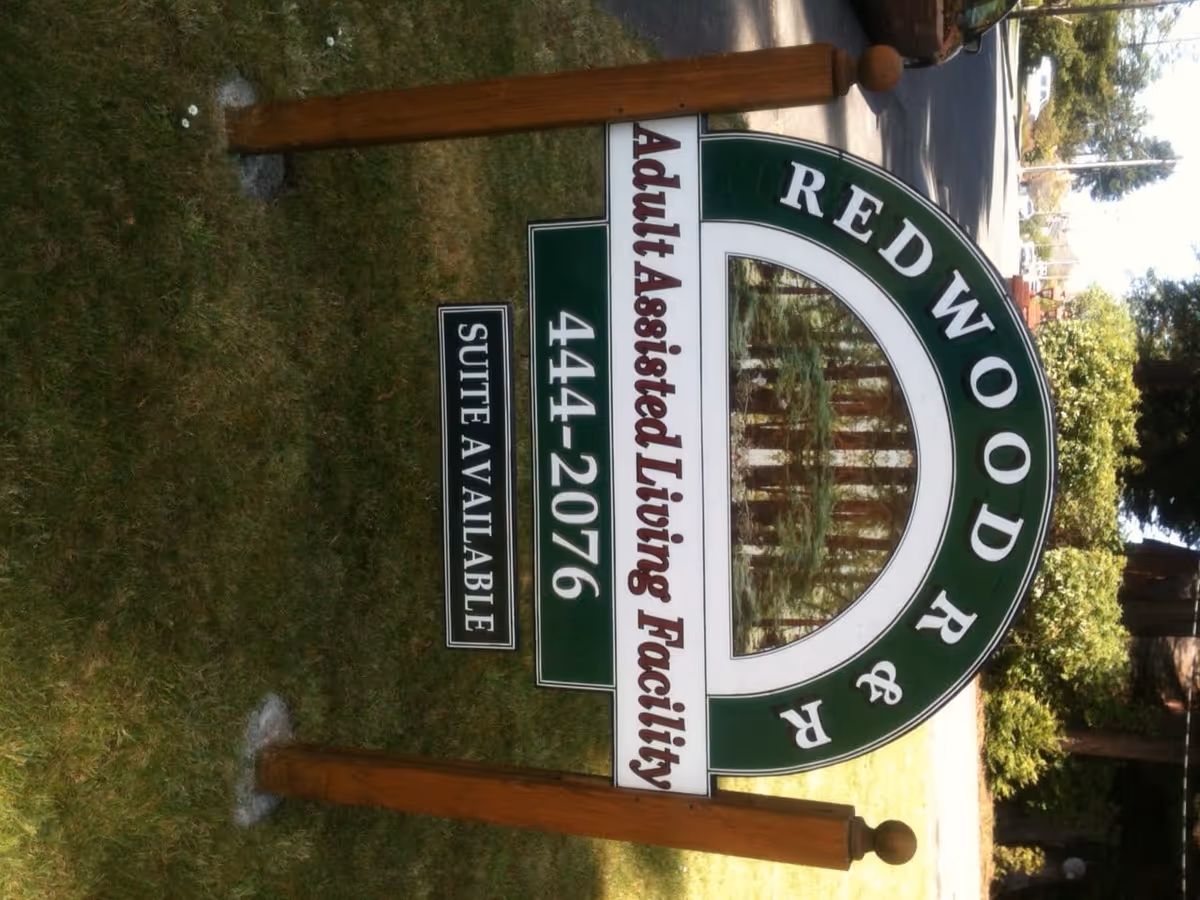 Outdoor sign for Redwood R & R Adult Assisted Living Facility with a phone number 444-2076 and a note that suites are available, set on a grassy area with trees and a street in the background.