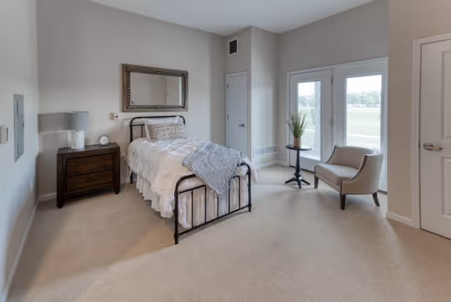 A bright and spacious bedroom with a single bed featuring a black metal frame, white bedding, and a gray throw blanket. Next to the bed is a dark wooden nightstand with a lamp and a clock. A large decorative mirror hangs on the wall above the bed. The room has beige carpet flooring, light gray walls, and a corner with a small round table holding a plant and a beige upholstered chair near glass doors that lead outside.