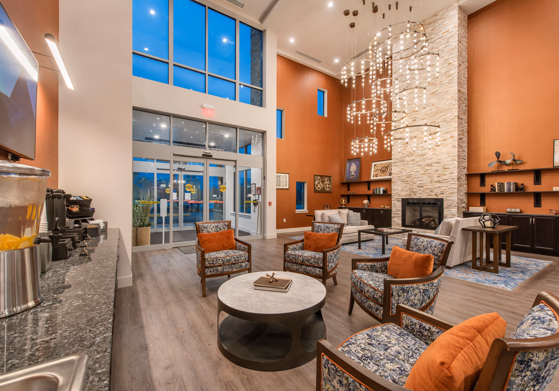 A spacious and modern senior living facility common area with high ceilings and large windows. The room features a seating arrangement with patterned armchairs and orange cushions around a round marble coffee table. There is a stone fireplace on the right wall with decorative shelves and artwork. A contemporary chandelier with multiple circular light fixtures hangs from the ceiling. On the left side, there is a countertop with a beverage dispenser and coffee machines. The entrance door with glass panels is visible in the background.