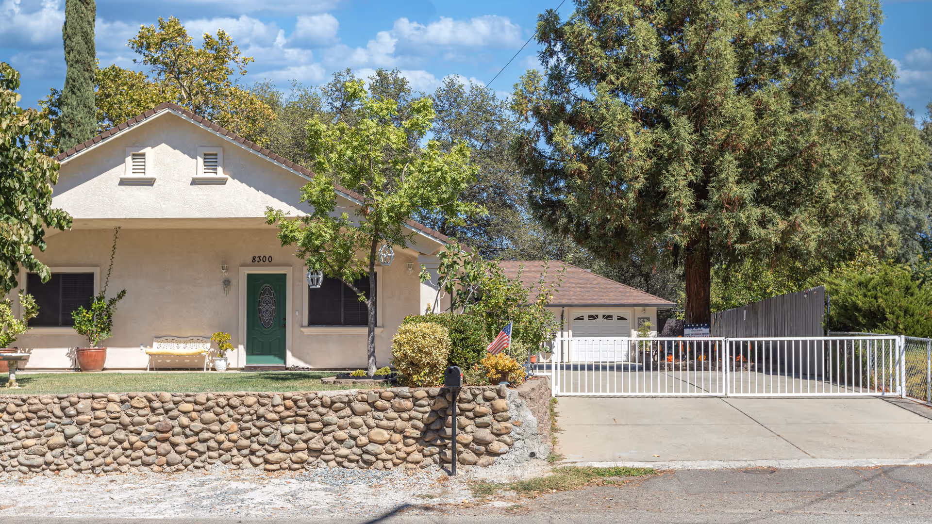 Exterior view of a single-story residential-style building with a green front door, two windows, and a stone retaining wall in front. There is a driveway with a white gate leading to a garage, surrounded by trees and shrubs under a blue sky with some clouds.