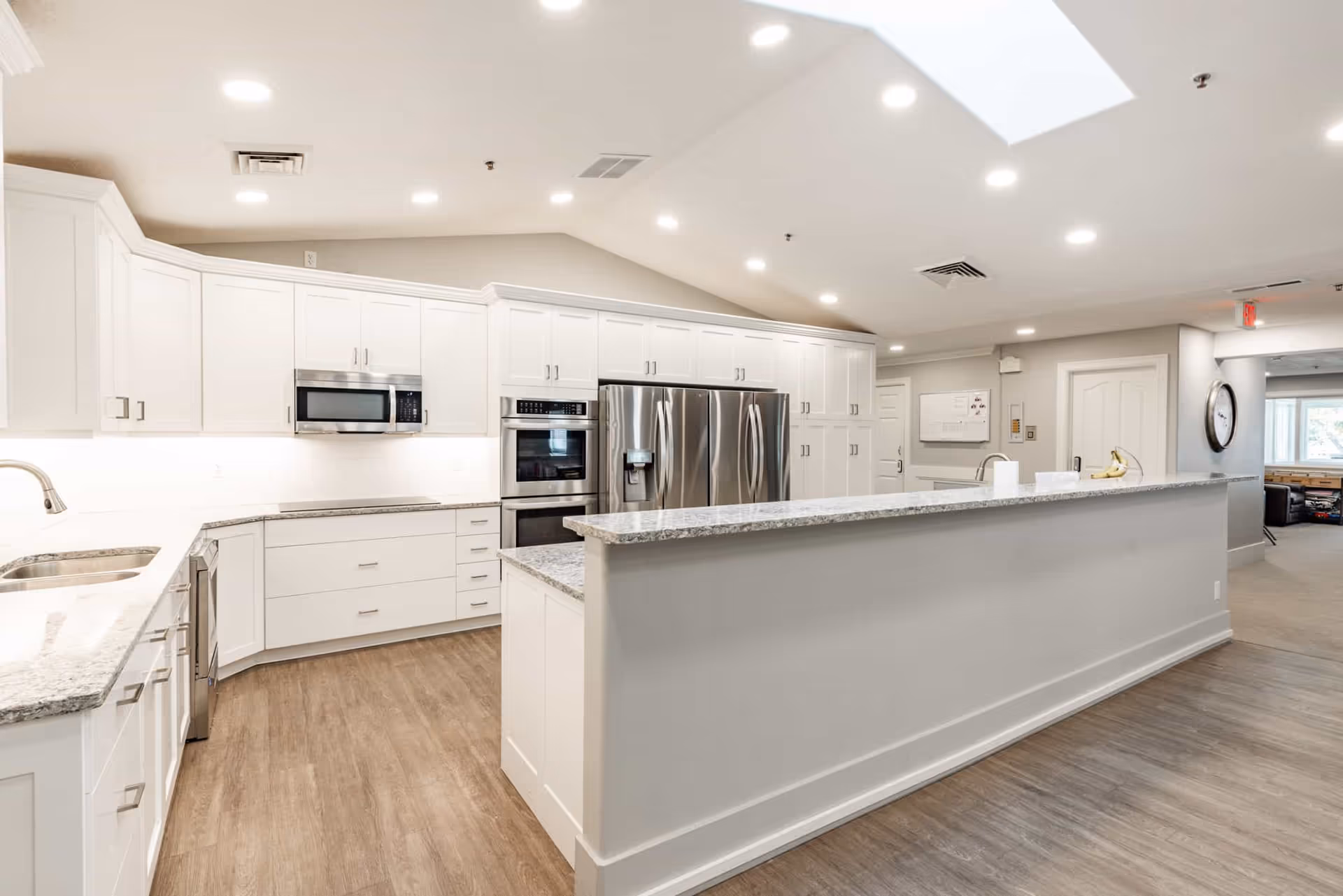 Bright modern communal kitchen with white cabinetry, a long granite-topped island, stainless steel appliances, and wood-look flooring.