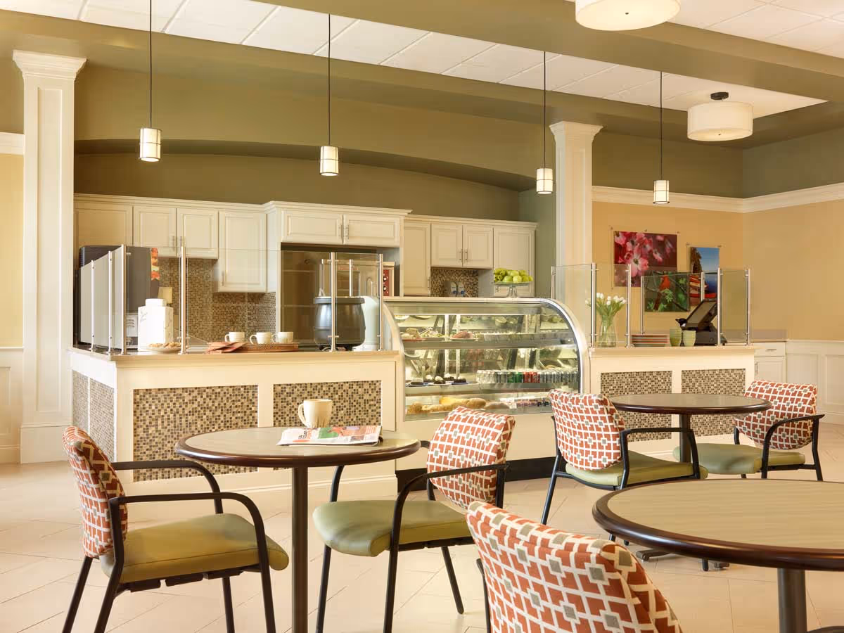 A bright and clean dining area with round tables and cushioned chairs featuring a geometric pattern. In the background, there is a counter with a glass display case containing various food items, white cabinetry, and pendant lights hanging from the ceiling. The walls are painted in soft green and beige tones with decorative artwork and flowers on the counter.