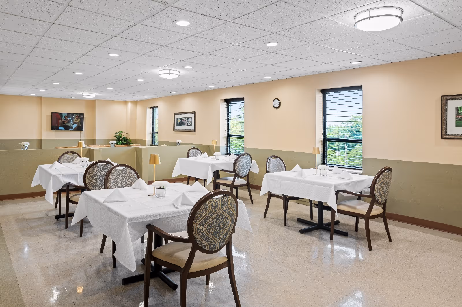 A clean and well-lit dining room with several tables covered in white tablecloths, each set with napkins and small lamps. The room has beige walls with a green lower half, windows with blinds, framed artwork, and a wall clock. There are patterned chairs around the tables and a television mounted on one wall.