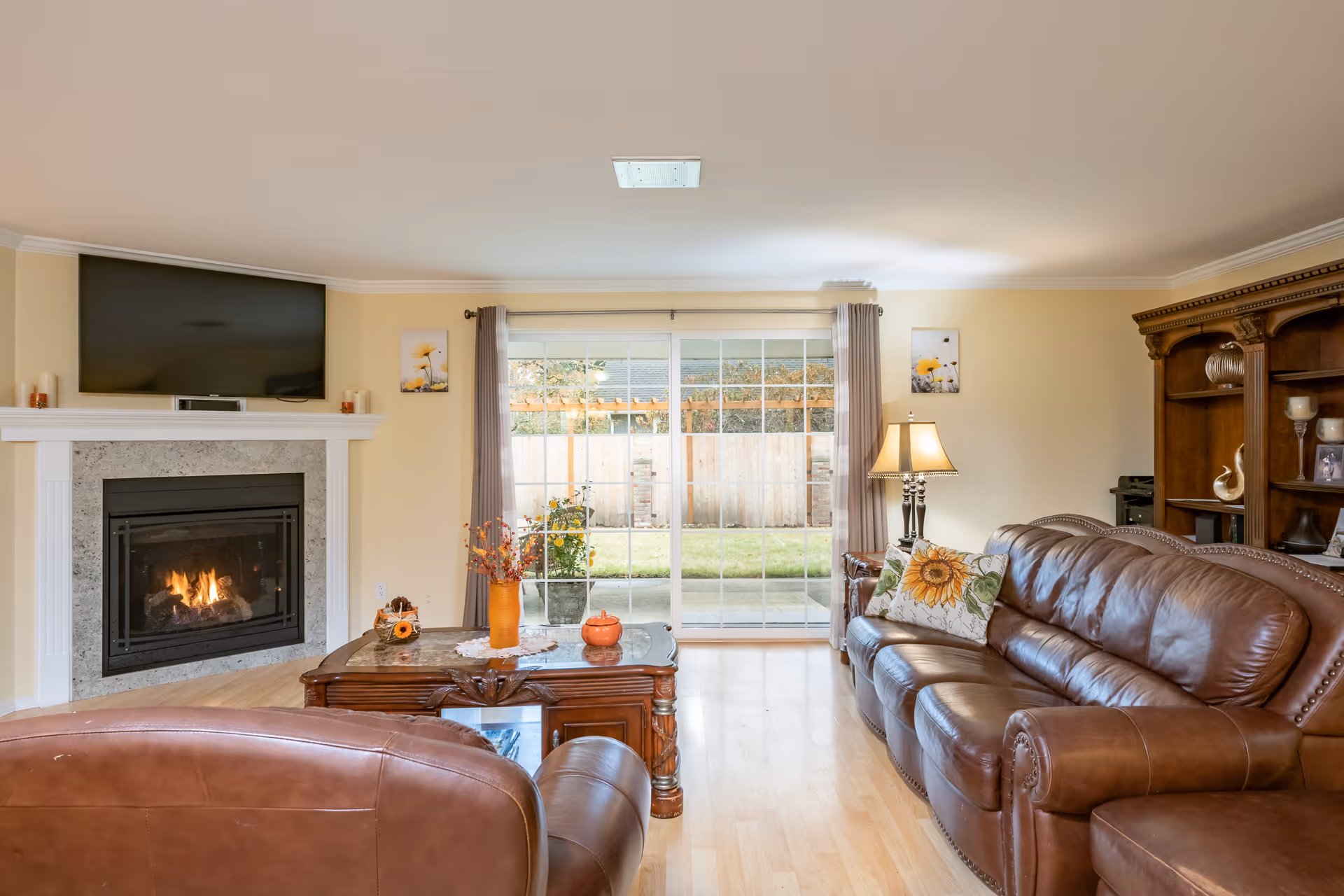 A cozy living room with a brown leather sofa and armchair, a wooden coffee table with decorative items, a lit fireplace with a mounted TV above it, and a large sliding glass door leading to a fenced backyard. The room has light-colored walls and wooden flooring, with a wooden shelving unit on the right side and a table lamp next to the sofa.