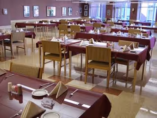 A spacious dining room with multiple tables covered in burgundy tablecloths, each set with plates, napkins folded in a triangular shape, and silverware. Wooden chairs are arranged around the tables, and the room has large windows allowing natural light to enter. The walls are decorated with framed pictures.