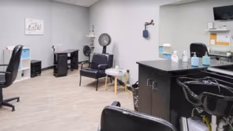 Interior view of a senior living facility room set up as a hair salon with black salon chairs, a hair dryer, a mirror, and shelves with hair care products. The room has light gray walls and light wood flooring.