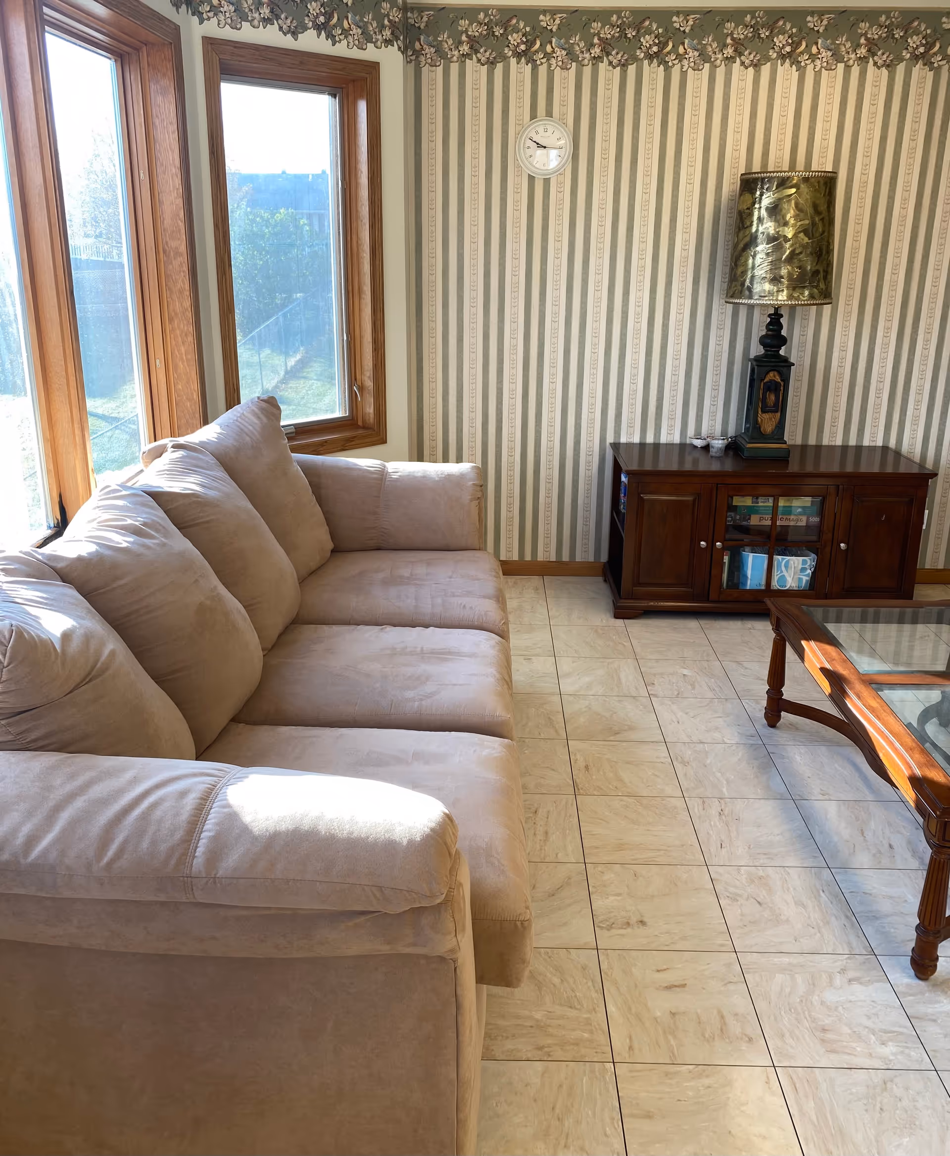 Sunlit living room with a beige sofa by large windows, striped wallpaper, a wooden cabinet with a lamp, and a glass-top coffee table on a tiled floor.