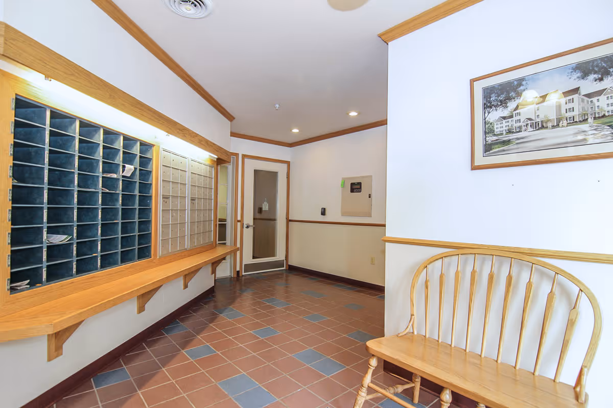 Interior view of a senior living facility mailroom with multiple mail slots and a row of locked mailboxes on the wall. There is a wooden bench on the right side beneath a framed picture, and the floor is tiled with a pattern of brown and blue tiles. The walls are white with wooden trim, and a door is visible at the end of the room.