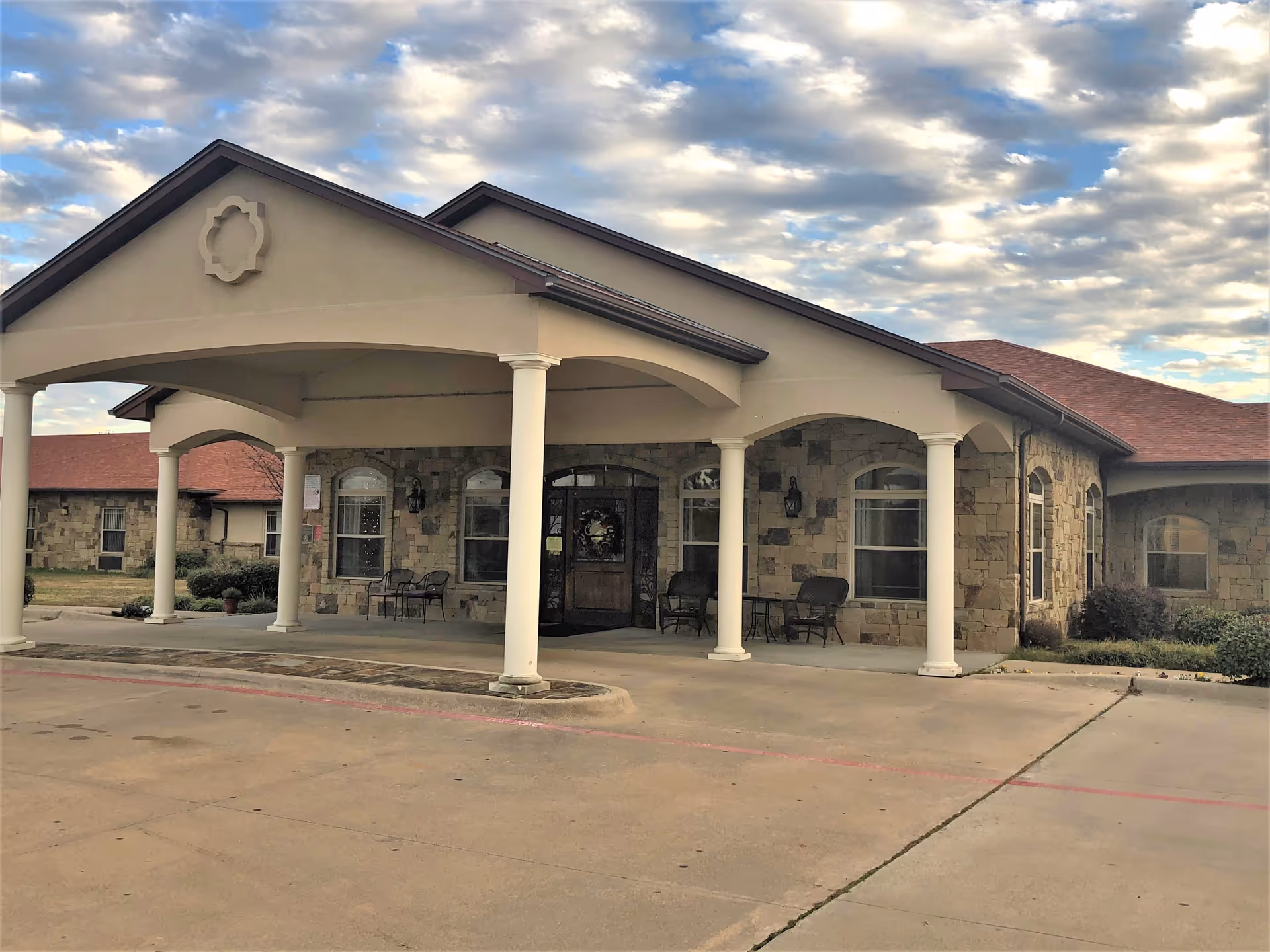 Front exterior view of a single-story building with stone walls and a covered entrance supported by white columns. There are chairs and small tables near the entrance, and the sky is partly cloudy.