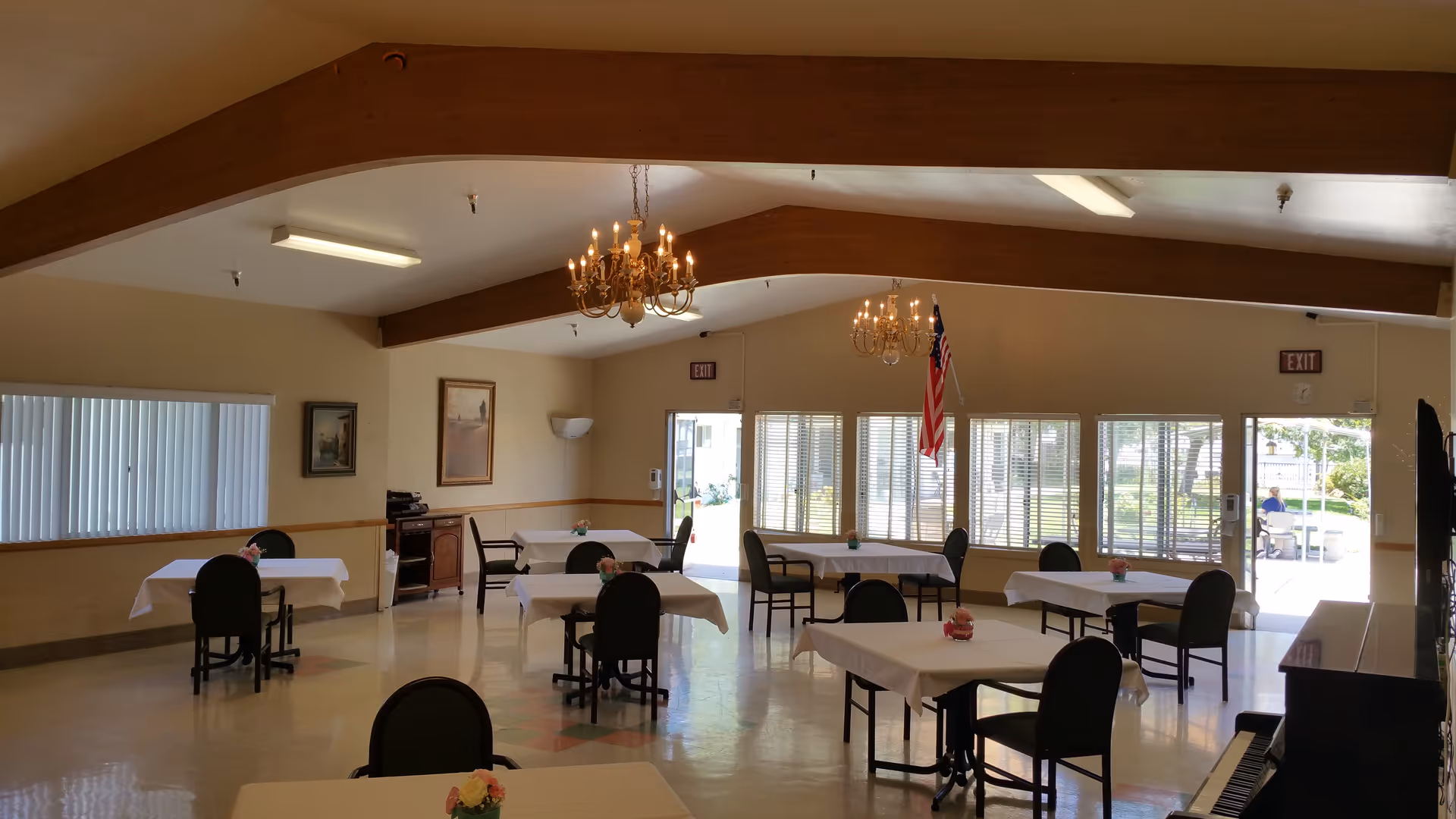Spacious dining/activity room with multiple tables and chairs, chandeliers, large windows and an American flag by the exit.