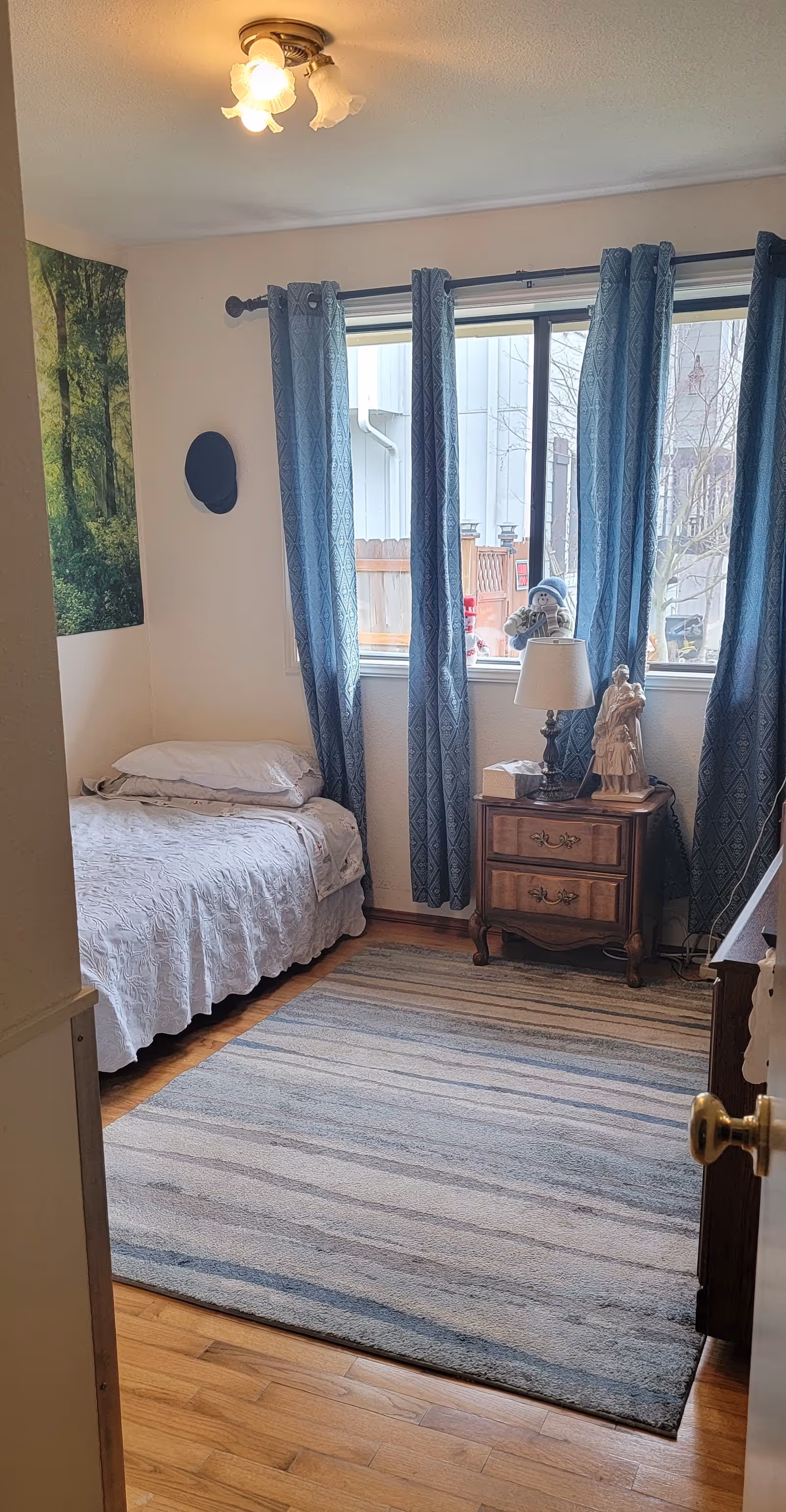 A small bedroom with a single bed covered in a white quilt, a wooden nightstand with a lamp and decorative figurines, blue patterned curtains on the window, a forest-themed wall poster, and a striped area rug on a wooden floor.
