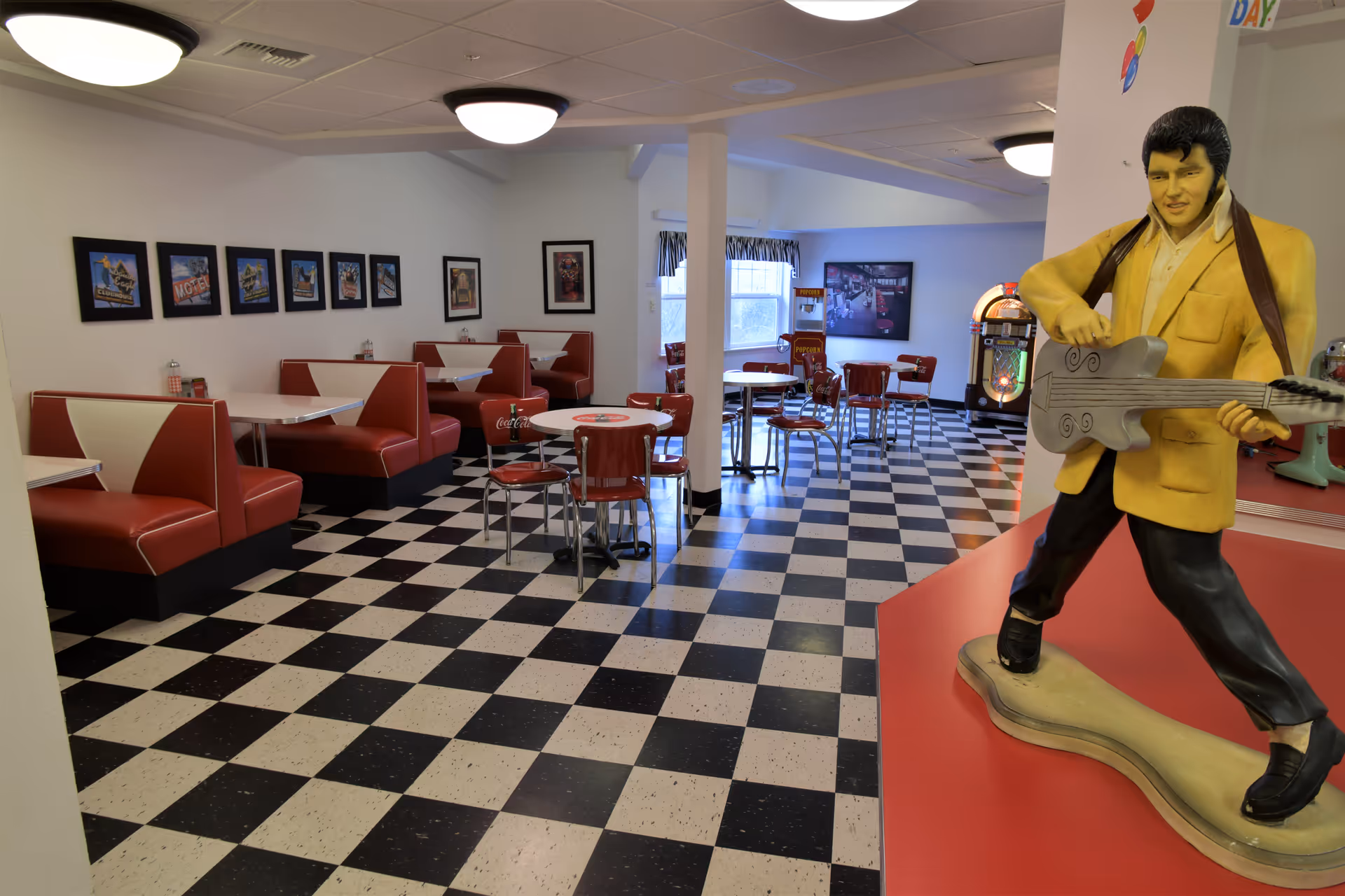 Interior of a retro-style dining area with black and white checkered floor tiles, red and white booth seating along the left wall, and round tables with red chairs in the center and back. On the right side, there is a statue of a man playing a guitar, a jukebox, and vintage decor. The walls have framed pictures and windows with curtains.