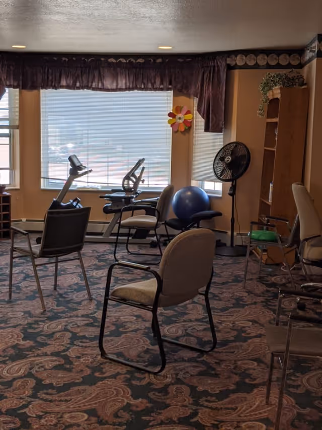 A room with several chairs arranged in a circle on a patterned carpet. There is exercise equipment including a stationary bike and a large blue exercise ball near the windows with blinds. A standing fan and a wooden bookshelf with some items on it are also visible. The room has beige walls and a valance over the windows.