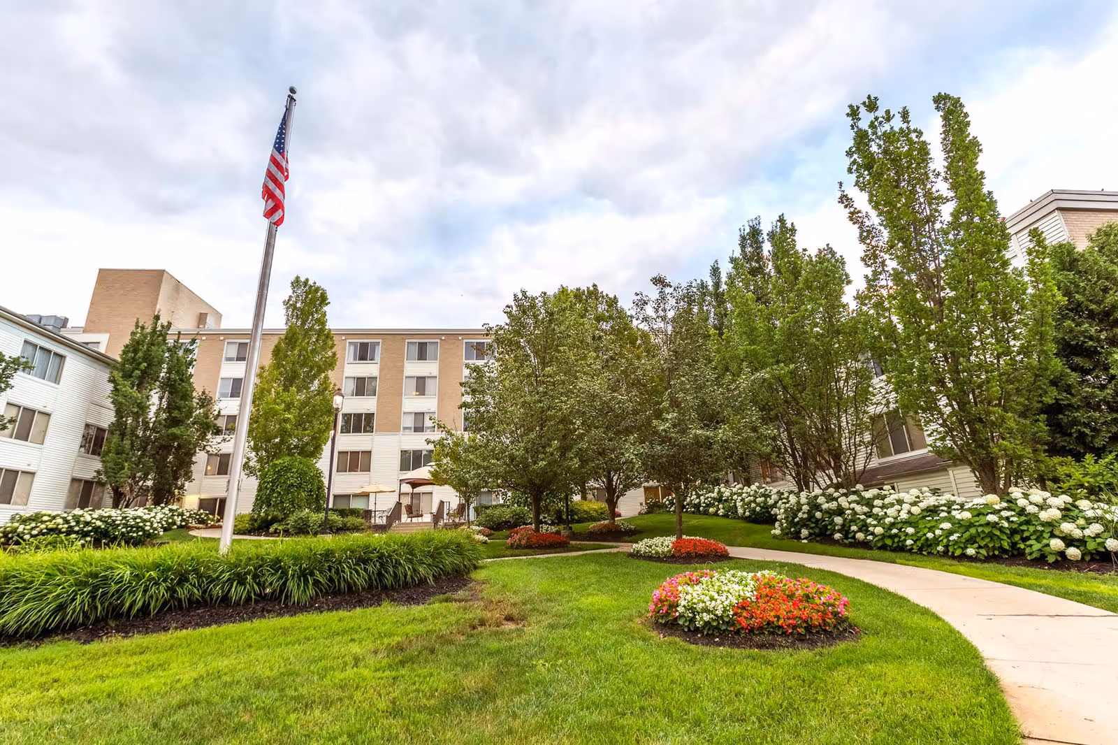 Outdoor garden area at Independence Village of Plymouth with a well-maintained lawn, flower beds, trees, a curved walkway, and an American flag on a flagpole. The background shows a multi-story residential building under a partly cloudy sky.